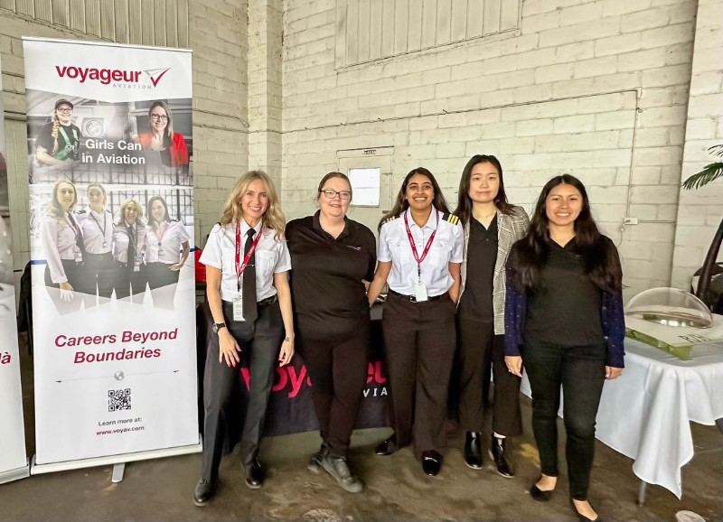 A group of women standing together at an event. They are in front of a banner for Voyageur Aviation, which includes the phrases Girls Can in Aviation and Careers Beyond Boundaries.
