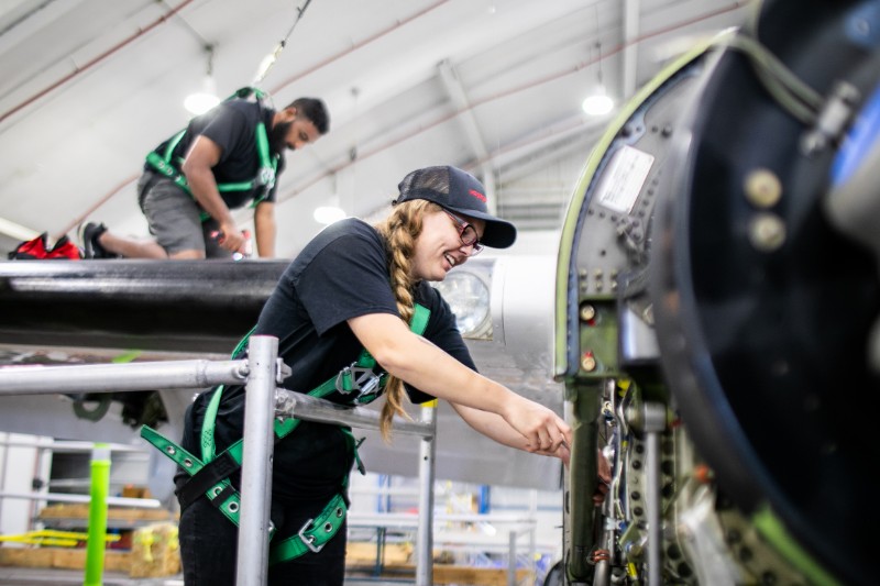Two individuals, a woman and a man, working on an aircraft. They are wearing safety harnesses and appear to be engaged in maintenance or repair work inside a hangar.