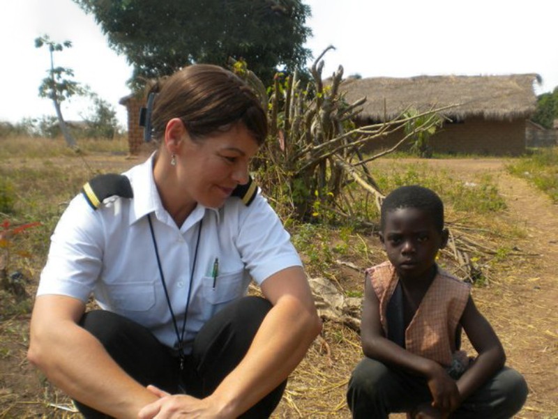 A female flight attendant in uniform, kneeling down and smiling at a young boy sitting beside her in a rural area. Behind them, there is a thatched-roof hut and some vegetation.