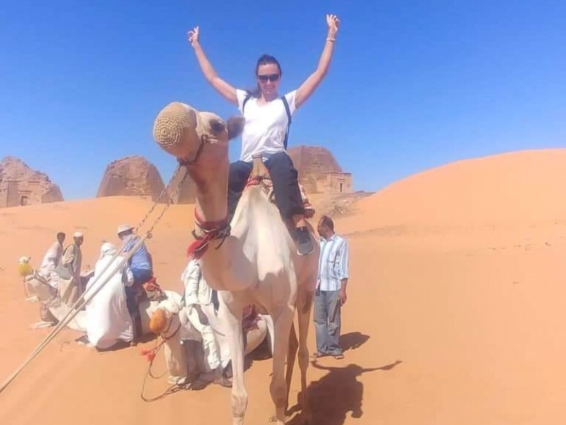 A person riding a camel in a desert landscape with hands raised in the air, smiling. In the background, there are small groups of people and ancient stone structures.