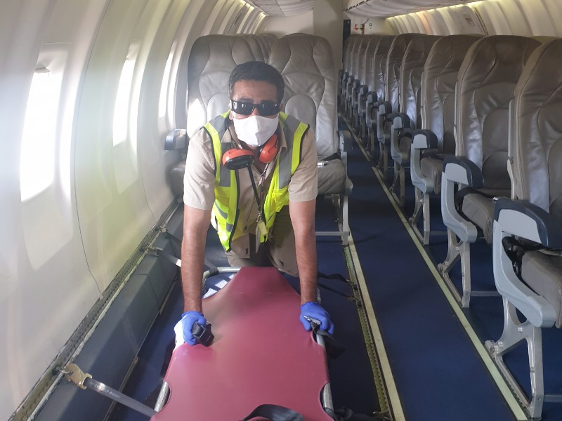 A person wearing a mask, gloves, and a high-visibility vest is seen inside an airplane, pushing a large red item along the aisle. The background shows rows of empty seats.